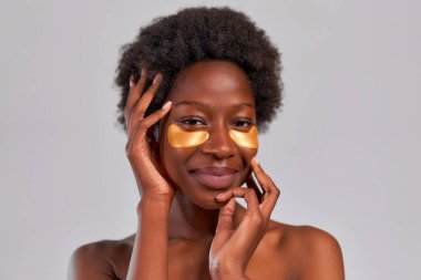 Gri arka planda izole edilmiş göz bantları kullanan afro saçlı ve mükemmel derecede sağlıklı cilde sahip Afro-Amerikalı genç bir kadın. Cilt bakımı ve kaplıca tedavisi.