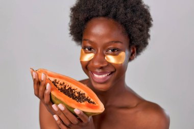 Afro saçlı, pürüzsüz, sağlıklı ciltli, göz bandı takan ve gri arka planda papaya tutan Afro-Amerikan genç bir kadın. Cilt bakımı ve kaplıca tedavisi.