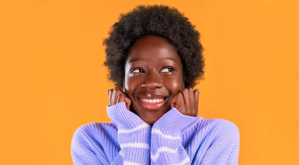 身穿蓝色毛衣 背景明亮橙色的非洲裔美国年轻女子留着一头非洲式的头发 她把拳头深深地插在下巴下 — 图库照片