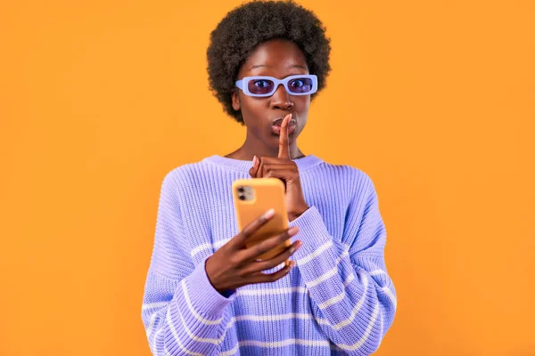 アフリカ系アメリカ人の若いです女性とともにアフロヘアスタイリングで立っています青いセーターと眼鏡で明るいオレンジの背景に携帯電話を使用して静かに指を彼女の口に置くジェスチャーを示しています — ストック写真