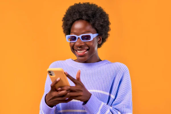 アフリカ系アメリカ人の若い女性で 青いセーターと眼鏡に身を包んだアフロヘアスタイルの女性は 携帯電話を使用して明るいオレンジ色の背景にあり 陽気に笑っています — ストック写真