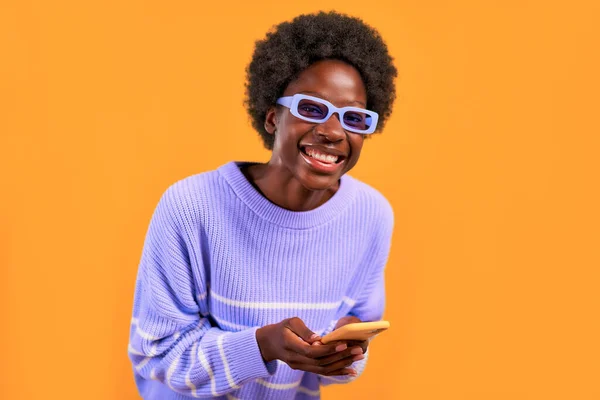 アフリカ系アメリカ人の若い女性で 青いセーターと眼鏡に身を包んだアフロヘアスタイルの女性は 携帯電話を使用して明るいオレンジ色の背景にあり 陽気に笑っています — ストック写真