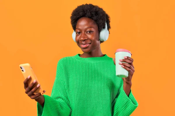 アフリカ系アメリカ人の若いですアフロの髪のスタイリングは コーヒーと携帯電話を使用してヘッドフォンで明るいオレンジの背景に緑のセーターに立って — ストック写真