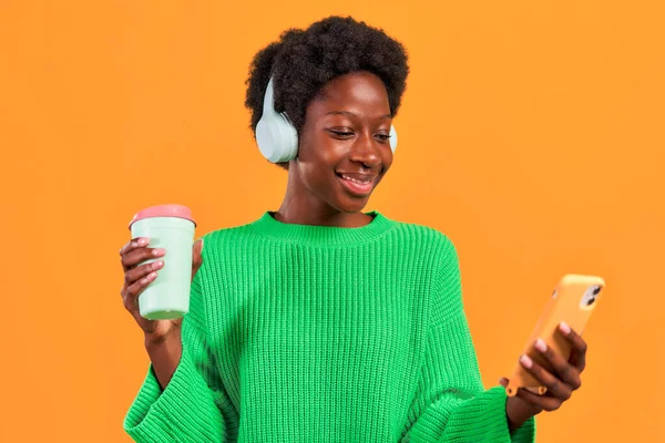 アフリカ系アメリカ人の若いですアフロの髪のスタイリングは コーヒーと携帯電話を使用してヘッドフォンで明るいオレンジの背景に緑のセーターに立って — ストック写真
