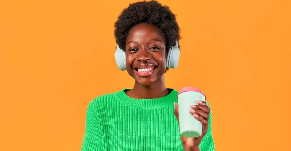 アフリカ系アメリカ人の若いです女性とともにアフロヘアスタイリングスタンディングで緑のセーターで明るいオレンジの背景にヘッドフォンとコーヒーを飲む — ストック写真