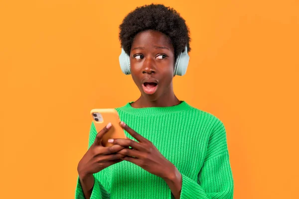 アフリカ系アメリカ人の若いです女性とともにアフロヘアスタイリング立っています緑のセーターで明るいオレンジの背景にヘッドフォンを身に着けていると携帯電話を使用して — ストック写真