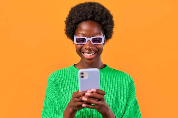アフリカ系アメリカ人の若いです女性とともにアフロヘアスタイリング立っています緑のセーターで明るいオレンジの背景に眼鏡をかけ 携帯電話を使用して — ストック写真