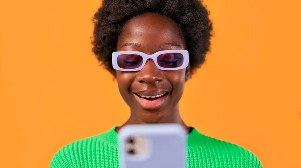 アフリカ系アメリカ人の若いです女性とともにアフロヘアスタイリング立っています緑のセーターで明るいオレンジの背景に眼鏡をかけ 携帯電話を使用して — ストック写真
