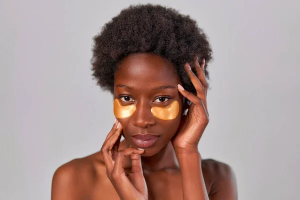 African American young woman with afro hair and perfect even healthy skin using eye patches isolated on gray background. Skin care and spa treatments.