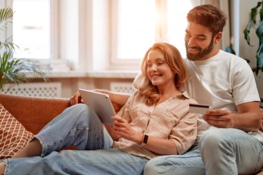 Tablet ve kredi kartıyla kanepede uzanan güzel yetişkin bir çift evdeki oturma odasında online alışveriş yapıyor, sohbet ediyor, romantik bir şekilde vakit geçiriyorlar..