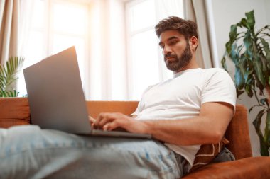 Pencerenin yanındaki kanepede dizüstü bilgisayarla oturan yakışıklı sakallı adam, online olarak çalışıyor, alışveriş yapıyor ya da evdeki oturma odasında sohbet ediyor..