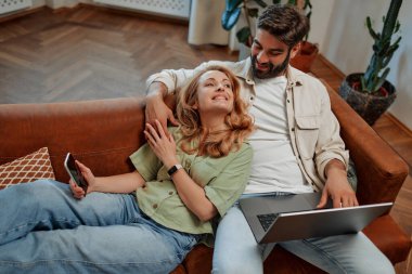 Yetişkin bir çift evdeki oturma odasında dizüstü bilgisayar ve telefonla kanepede uzanıyor, online çalışıyor, dinleniyor, sarılıyor ve birlikte zaman geçiriyorlar..