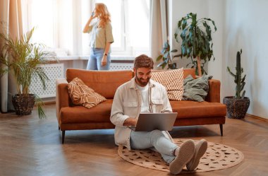 Kanepenin yanında dizüstü bilgisayarı olan yakışıklı sakallı bir adam arka planda telefonla konuşan bir kadın. Çift hafta sonunu birlikte geçirecek..