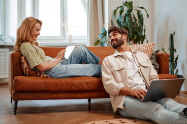 Evde dizüstü bilgisayar ve tabletle oturan güzel yetişkin bir çift online çalışıyor, alışveriş yapıyor ya da internette sohbet ediyorlar..