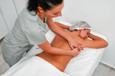 Rahatlamış genç bir kadının masaj masasına yüzüstü uzanması ve profesyonel masör tarafından spa salonunda ya da sağlık merkezinde yapılan vücut masajının keyfini çıkarması.