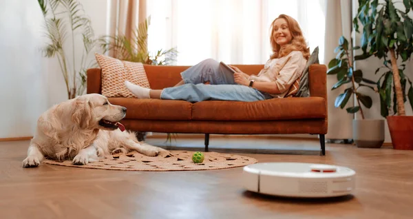 A beautiful blonde woman is sitting on a sofa with a tablet in the living room at home along with a labrador dog, in the foreground a robot vacuum cleaner cleans the house.