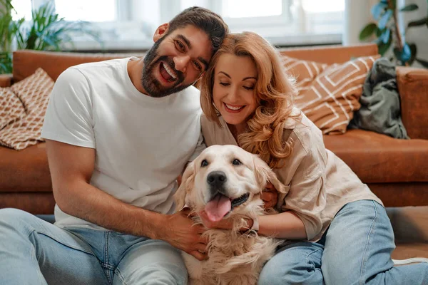 Young couple playing and hugging their pet labrador dog while sitting on the sofa by the window in the living room at home, having fun together.