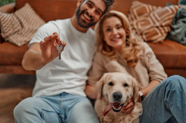 Moving buying a new apartment, house. Adult couple in love with their labrador dog, holding house keys, sitting on the floor near the sofa in the living room in the new apartment.