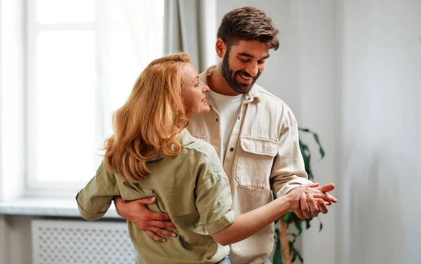 stock image An adult couple in love dancing merrily in the living room against the background of the sofa and the window at home, having fun and romantic time together.