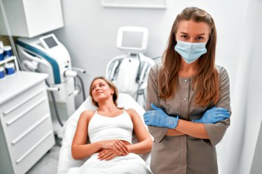 Bir kaplıca salonundaki kanepede uzanmış genç bir kadın ve kollarını kavuşturmuş tıbbi koruyucu maskeli bir güzellik uzmanı. Vücut ve yüz bakımı. Kozmetoloji, kaldırma, epilasyon kavramı.