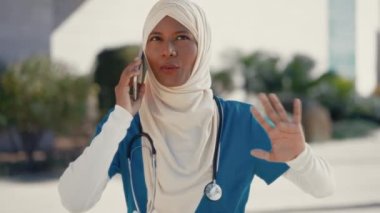 Üniformalı, tesettürlü, steteskoplu genç Müslüman bir kadın hastane binasının yanında caddede dikilirken telefonla konuşuyor. İlaç ve sağlık hizmetleri.