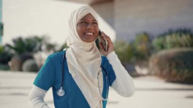Üniformalı, tesettürlü, steteskoplu genç Müslüman bir kadın hastane binasının yanında caddede dikilirken telefonla konuşuyor. İlaç ve sağlık hizmetleri.