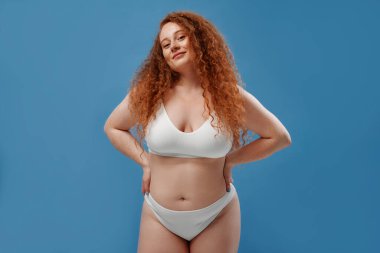 Genç, güzel, kızıl saçlı, kilolu ya da tombul bir kadın doğal bedenini kutluyor. Mavi arka planda beyaz iç çamaşırlı güzel bir kadın model. Minimum makyajlı yaşam tarzı portresi