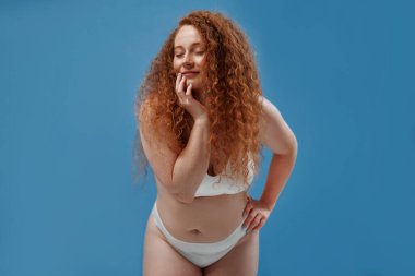 Genç, güzel, kızıl saçlı, kilolu ya da tombul bir kadın doğal bedenini kutluyor. Mavi arka planda beyaz iç çamaşırlı güzel bir kadın model. Minimum makyajlı yaşam tarzı portresi