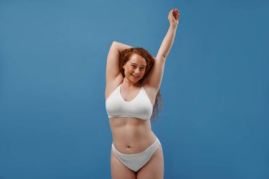 Genç, güzel, kızıl saçlı, kilolu ya da tombul bir kadın doğal bedenini kutluyor. Mavi arka planda beyaz iç çamaşırlı güzel bir kadın model. Minimum makyajlı yaşam tarzı portresi
