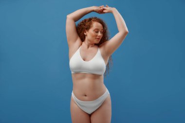 Genç, güzel, kızıl saçlı, kilolu ya da tombul bir kadın doğal bedenini kutluyor. Mavi arka planda beyaz iç çamaşırlı güzel bir kadın model. Minimum makyajlı yaşam tarzı portresi