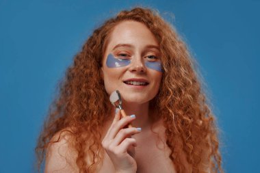 Stüdyo dermatolojisi, sağlık hizmetleri veya cilt bakımı için kadın boyu güzellik, göz bandı ve yüz silindiri. Mavi arka planda kızıl saçlı, tombul bir kadın. Vücut pozitif.