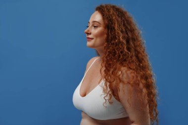 Genç, güzel, kızıl saçlı, kilolu ya da tombul bir kadın doğal bedenini kutluyor. Mavi arka planda beyaz iç çamaşırlı güzel bir kadın model. Minimum makyajlı yaşam tarzı portresi