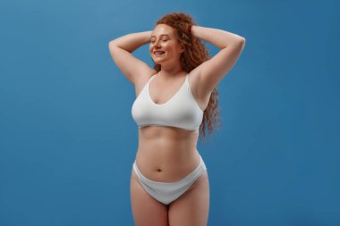 Genç, güzel, kızıl saçlı, kilolu ya da tombul bir kadın doğal bedenini kutluyor. Mavi arka planda beyaz iç çamaşırlı güzel bir kadın model. Minimum makyajlı yaşam tarzı portresi