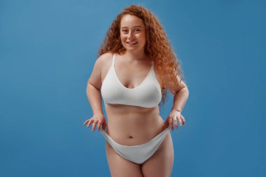 Genç, güzel, kızıl saçlı, kilolu ya da tombul bir kadın doğal bedenini kutluyor. Mavi arka planda beyaz iç çamaşırlı güzel bir kadın model. Minimum makyajlı yaşam tarzı portresi