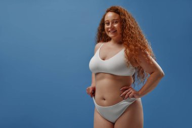 Genç, güzel, kızıl saçlı, kilolu ya da tombul bir kadın doğal bedenini kutluyor. Mavi arka planda beyaz iç çamaşırlı güzel bir kadın model. Minimum makyajlı yaşam tarzı portresi