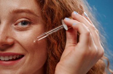 Çilli, kızıl saçlı, kilolu bir kadın. Yüzüne kırışıklık önleyici nemlendirici serum sürüyor. Mavi arka planda izole edilmiş. Cilt bakımı, vücut pozitifliği ve kozmetoloji kavramı.