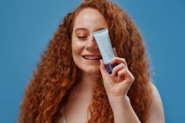 Çilli kızıl saçlı, kilolu, beyaz iç çamaşırlı, elinde fondöten kremi ya da mavi arka planda izole edilmiş kapatıcı tutan bir kadın. Cilt bakımı, vücut pozitifliği ve kozmetoloji kavramı.