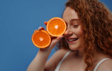 Çilli kızıl saçlı, iri yarı, beyaz iç çamaşırlı, mavi arka planda turuncu yarımlar tutan bir kadın. Yüz ve vücut bakımı, vücut pozitifliği ve kozmetoloji kavramı.