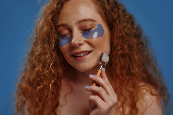 stock image Women's plus size beauty, eye patch and face roller for studio dermatology, healthcare or skincare. A red-haired plump woman on a blue background. Body positive.