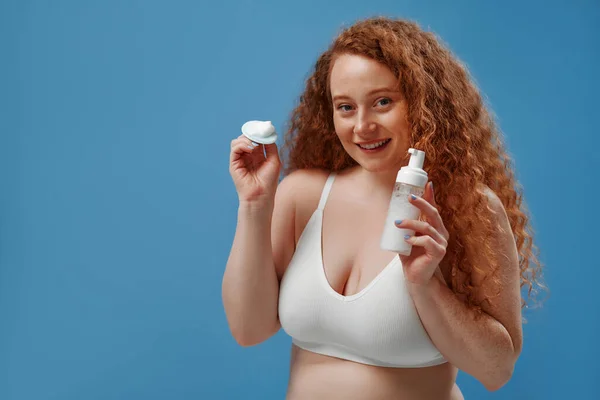 Freckled redhead plump plus size woman in white lingerie using facial cleanser and silicone brush isolated on blue background. The concept of skin care, body positive and cosmetology.