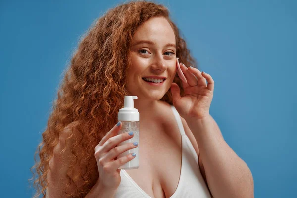 Freckled redhead plump plus size woman in white lingerie using facial cleanser and silicone brush isolated on blue background. The concept of skin care, body positive and cosmetology.