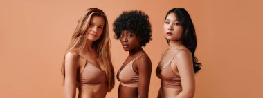 Bej iç çamaşırlı çok ırklı genç kadınlar bej arka planda izole edilmiş. Stüdyoda poz veren Afro-Amerikalı, Kafkas ve Asyalı kadınlar. Yüz bakımı, kozmetoloji kavramı.