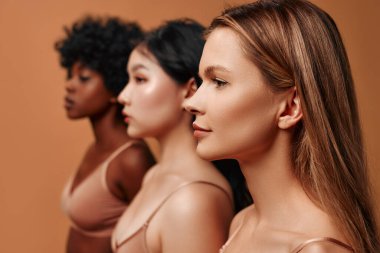 Bej iç çamaşırlı çok ırklı genç kadınlar bej arka planda izole edilmiş. Stüdyoda poz veren Afro-Amerikalı, Kafkas ve Asyalı kadınlar. Yüz bakımı, kozmetoloji kavramı.