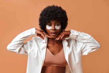 Güzel bir Afro-Amerikalı kadın, iç çamaşırlarıyla nemlendirici göz bantları kullanıyor ve stüdyoda bej arka planda poz veriyor. Kozmetik ve kaplıca cilt bakımı.