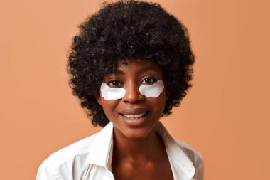 Güzel bir Afro-Amerikalı kadın, iç çamaşırlarıyla nemlendirici göz bantları kullanıyor ve stüdyoda bej arka planda poz veriyor. Kozmetik ve kaplıca cilt bakımı.