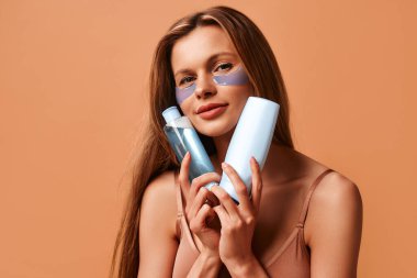 İç çamaşırlı güzel, zayıf bir kadın göz bandı kullanıyor ve bej bir arka planda izole edilmiş güzellik ürünleri şişeleri tutuyor. Kozmetoloji, cilt bakımı ve kaplıca tedavileri.