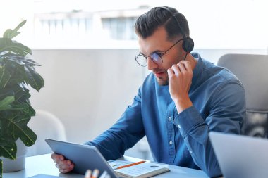 Ekibiyle birlikte çağrı merkezi çalışanı. Erkek işadamı serbest çalışan, birlikte çalışan, modern bir ofis. Kulaklık ve tabletle çalışan genç bir çalışan..