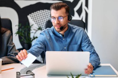 Ekibiyle birlikte çağrı merkezi çalışanı. Erkek işadamı serbest çalışan, birlikte çalışan, modern bir ofis. Kulaklık ve dizüstü bilgisayarla çalışan genç bir çalışan.