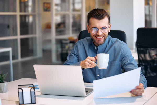 Young bearded handsome businessman in glasses and shirt sitting in a modern office with a laptop with a cup of coffee. Successful hipster team in coworking. Freelancers.