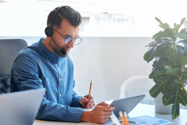 Call center employee accompanied by his team. Male businessman freelancer work in coworking, modern office. Young employee working with headset and tablet.